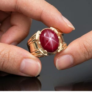 May include: A gold ring featuring a large, oval-shaped ruby gemstone with a six-ray star effect. The ring has a thick band and decorative accents on the sides. The ruby is a deep red color.