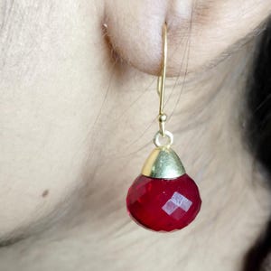 Puede incluir: Un solo pendiente dorado con una gema roja facetada en forma de lágrima. El pendiente cuelga de un gancho dorado, con una tapa dorada pulida en la parte superior de la gema. La gema tiene un corte brillante.