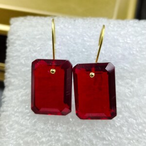 May include: A pair of red rectangular earrings with gold-colored hooks. Each earring features a faceted, ruby-red gemstone with a small gold accent near the top. The earrings are displayed against a white, textured background.