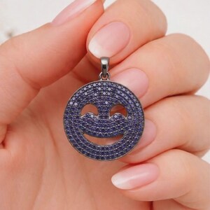 Puede incluir: Un colgante redondo con un diseño de carita sonriente. El colgante está cubierto de pequeñas piedras azul oscuro, creando un efecto brillante. El colgante está sostenido por una mano.