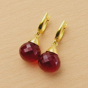 May include: A pair of gold-colored earrings with faceted, deep red teardrop-shaped gems. The earrings have a lever-back closure and a gold-colored cap that connects the gem to the earring hook. The gems have a multi-faceted surface.