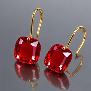 Pode incluir: Um par de brincos pendentes com pedras facetadas de um vermelho vibrante em corte quadrado, suspensas por ganchos curvos de tom dourado polido. As pedras têm um formato de almofada com cantos arredondados que refletem a luz em suas superfícies.