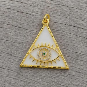 Op de afbeelding: Goudkleurige hanger in driehoekige vorm, met een witte achtergrond en een oogontwerp. Het oog is gedetailleerd met een cirkel van heldere stenen en een groene middensteen. De hanger heeft een lus voor een ketting.