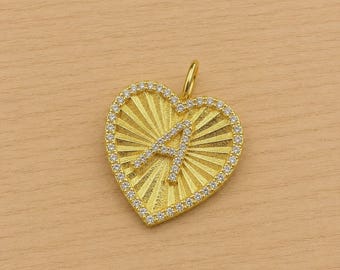 Puede incluir: Un colgante dorado en forma de corazón con un patrón texturizado de rayos de sol que irradian desde el centro. La letra A se muestra en el medio y todo el perímetro del corazón está bordeado por pequeñas piedras claras y brillantes.