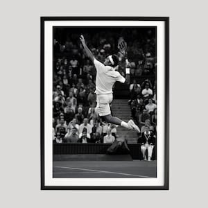May include: Black and white framed print of a tennis player mid-air, reaching for the ball with a racket. The player is wearing a white tennis outfit. The background shows a crowd of spectators.