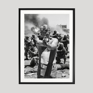May include: Black and white framed print of a shirtless man aiming a handgun, with a group of soldiers in the background. The image captures a moment of action, with smoke and debris visible.