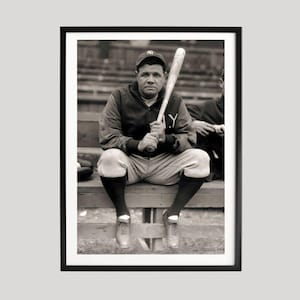 May include: Black and white framed photograph of a baseball player seated on a bench, holding a baseball bat. The player is wearing a baseball cap, uniform, and socks. The image is a vintage portrait.