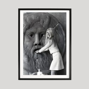 May include: Framed black and white photograph of a woman in a white outfit touching a stone sculpture of a face. The photograph is presented in a black frame.