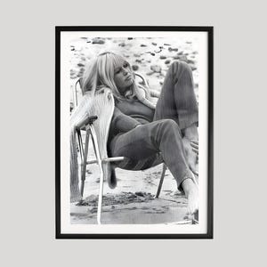 May include: Black and white framed photograph of a person seated in a chair on a beach. The person is wearing a sweater and trousers. The photograph is in a black frame.