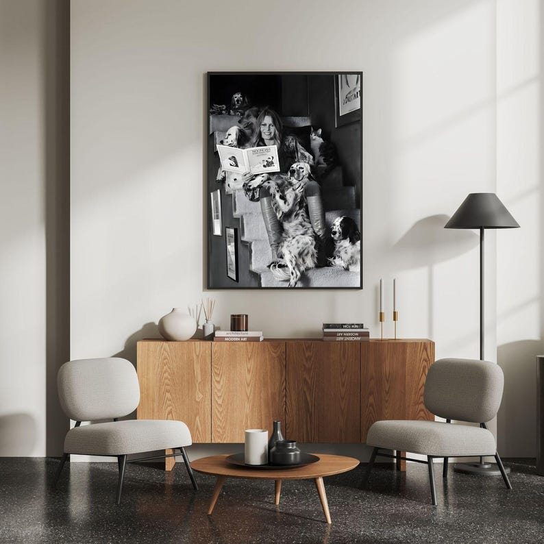 Puede incluir: Una impresi&oacute;n enmarcada en blanco y negro de una mujer leyendo un libro rodeada de perros. La impresi&oacute;n cuelga sobre un mueble de madera con una mesa de centro redonda y dos sillas grises. Tambi&eacute;n se ve una l&aacute;mpara de pie negra y velas.