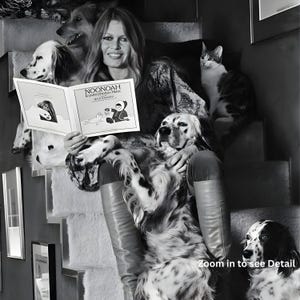 Puede incluir: Fotograf&iacute;a en blanco y negro de una mujer sentada en unas escaleras, rodeada de perros y un gato. Sostiene un libro titulado "NOONOAH le petit phoque blanc" con ilustraciones. La mujer sonr&iacute;e y las mascotas parecen relajadas.