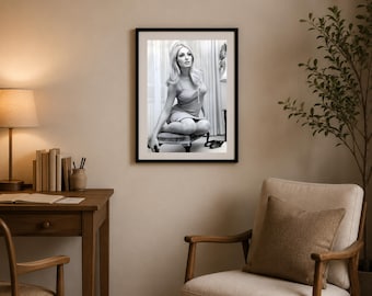 May include: A black and white framed wall art print featuring a portrait of a person with voluminous hair, wearing a sleeveless dress, sitting on a small stool. The person is positioned in front of curtains, with a pair of shoes visible on the floor.