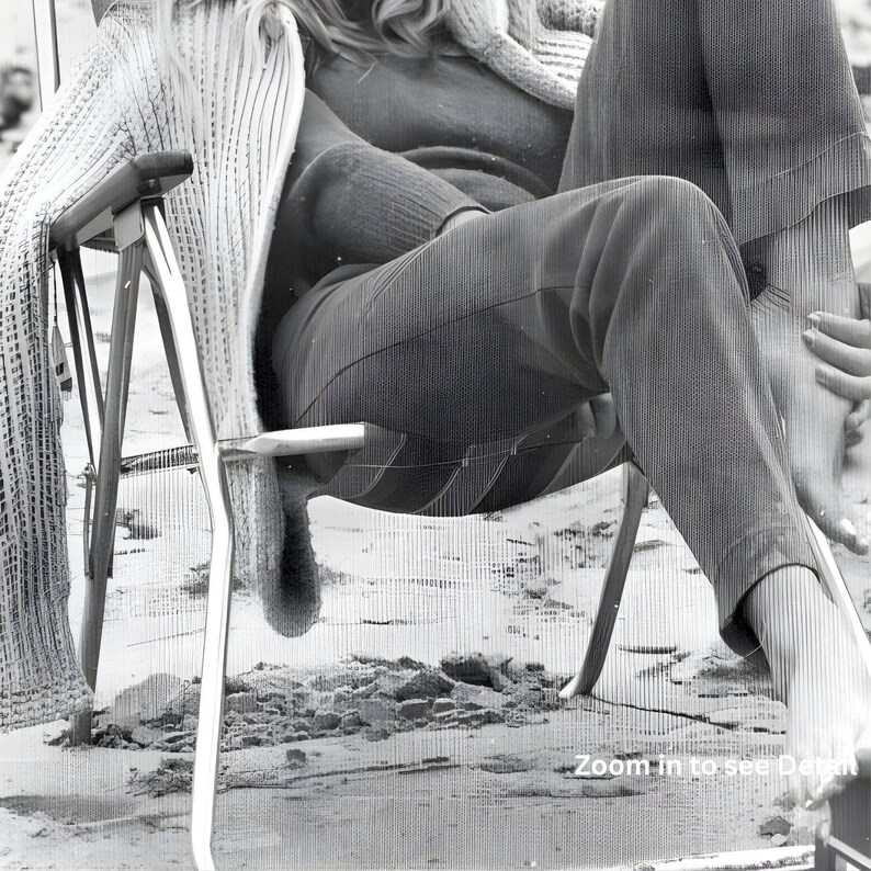 May include: A monochrome photograph showing a person seated in a folding chair on a beach. The person is wearing trousers, a sweater, and a striped cardigan. The chair has a metal frame and wooden armrests. A knitted blanket is draped over the chair.