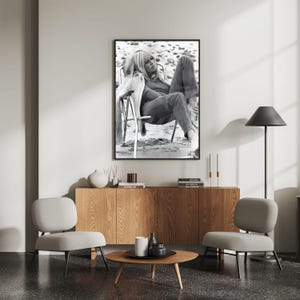 May include: A monochrome framed print of a woman seated on a beach, displayed above a wooden cabinet. The room features two grey chairs and a round coffee table. The decor is minimalist and stylish.
