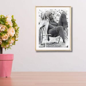 May include: A framed black and white photograph of a person sitting in a chair on a beach. The person is wearing a sweater and trousers. The photograph is in a gold frame and is displayed on a white wall.