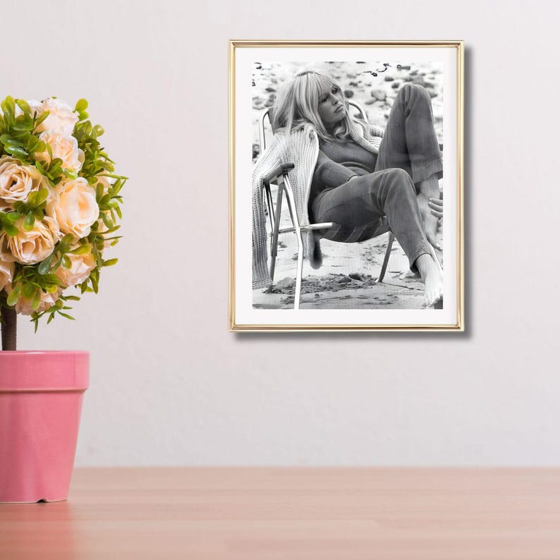 May include: A framed black and white photograph of a person sitting in a chair on a beach. The person is wearing a sweater and trousers. The photograph is in a gold frame and is displayed on a white wall.