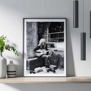 Pu&ograve; includere: Stampa incorniciata in bianco e nero di una donna che suona una chitarra acustica. La donna &egrave; seduta all'aperto, indossa una maglia scura a maniche lunghe e jeans. L'immagine &egrave; esposta su una mensola di legno con vasi decorativi e luci a sospensione.