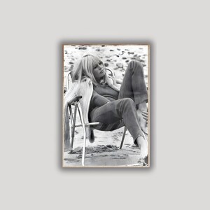 May include: Monochrome photograph of a person relaxing in a folding chair on a beach. The person is wearing a sweater and trousers. The image is framed in a wooden border.