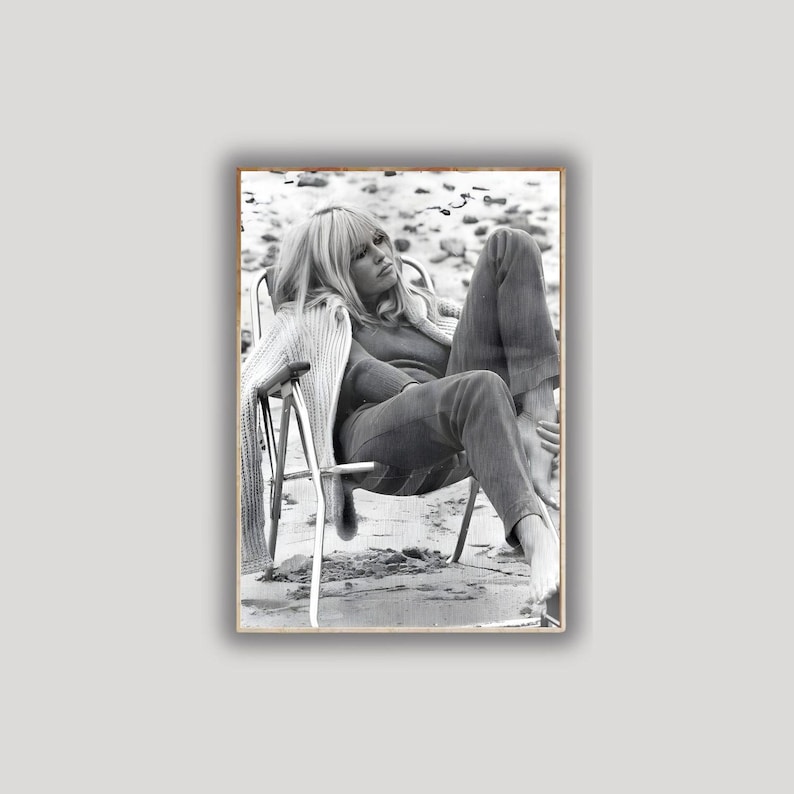 May include: Monochrome photograph of a person relaxing in a folding chair on a beach. The person is wearing a sweater and trousers. The image is framed in a wooden border.