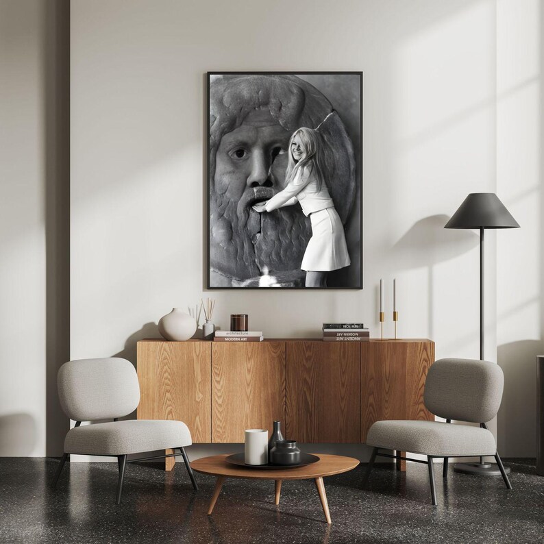 May include: A monochrome framed print featuring a woman and a stone sculpture of a face. The print is above a wooden cabinet with decor. Two grey chairs and a round coffee table are in the room.