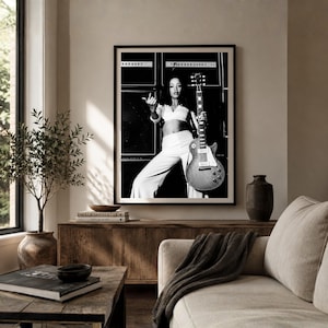 May include: A black and white framed wall art print featuring a person with long dark hair wearing a cropped top and wide-leg trousers, holding an electric guitar in front of a stack of large amplifiers. The print is displayed in a thin black frame.