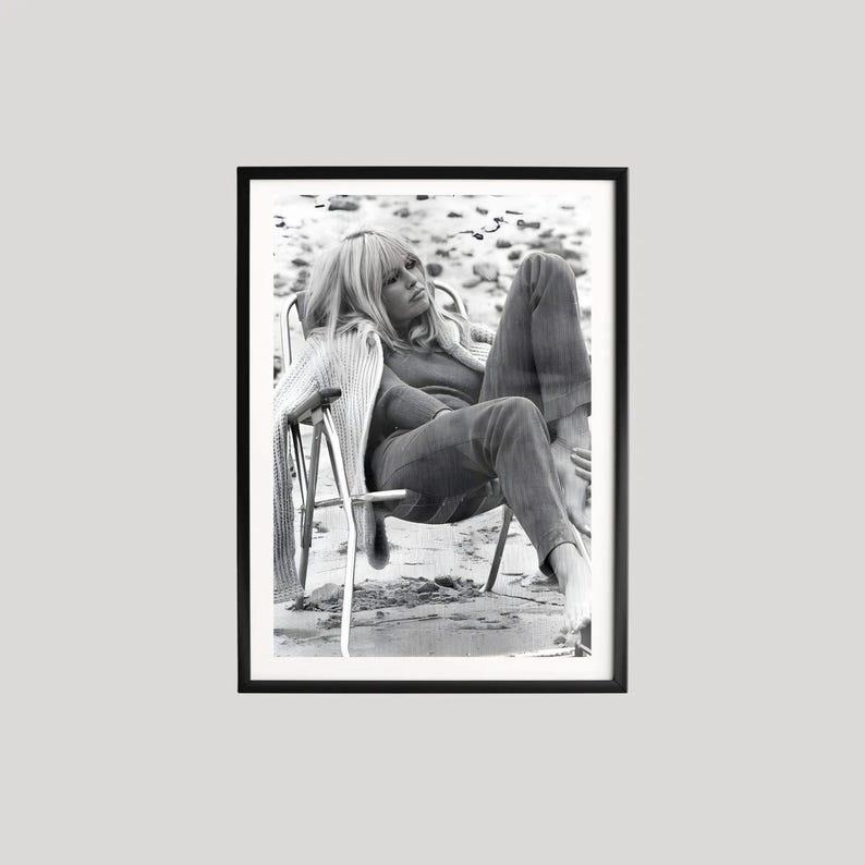 May include: Black and white framed photograph of a person seated in a chair on a beach. The person is wearing a sweater and trousers. The photograph is in a black frame.
