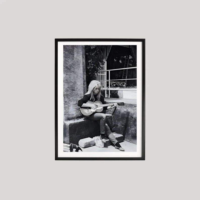 Pu&ograve; includere: Fotografia in bianco e nero incorniciata di una persona che suona una chitarra acustica. La persona &egrave; seduta su una struttura in pietra, indossa una maglia scura e jeans. La chitarra &egrave; marrone chiaro con un battipenna scuro.