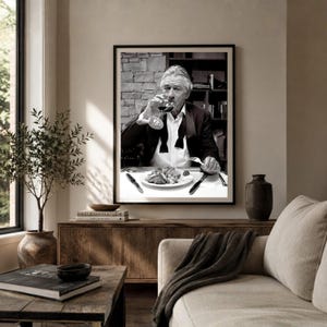 May include: A black and white framed wall art print featuring a man in a tuxedo jacket and bowtie sitting at a table. The man is holding a wine glass to his lips, with a plate of food, a fork, and a knife positioned in front of him.