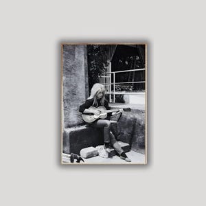 Pu&ograve; includere: Fotografia in bianco e nero di una donna che suona una chitarra acustica. &Egrave; seduta all'aperto, indossa una giacca scura e jeans. L'immagine ha un'estetica vintage, con un focus sulla musicista e il suo strumento.