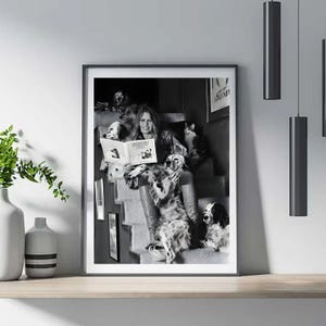 Puede incluir: Impresi&oacute;n enmarcada en blanco y negro que muestra a una mujer leyendo un libro titulado "NOONOW", rodeada de varios perros y un gato en una escalera. La imagen es un retrato.