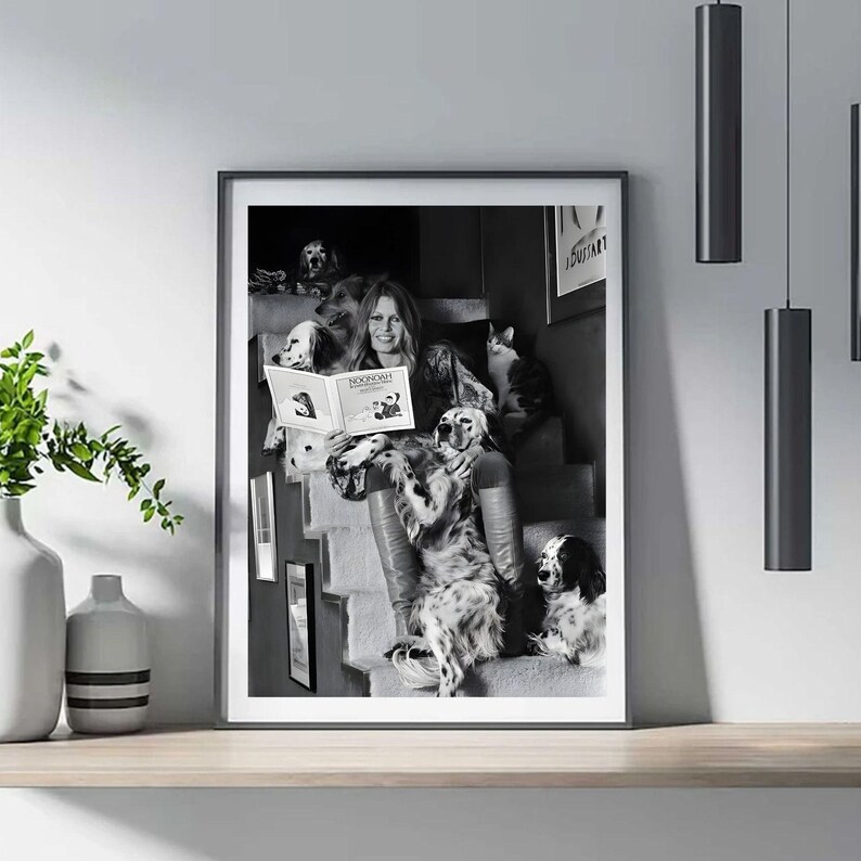 Puede incluir: Impresi&oacute;n enmarcada en blanco y negro que muestra a una mujer leyendo un libro titulado "NOONOW", rodeada de varios perros y un gato en una escalera. La imagen es un retrato.