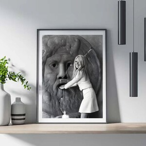May include: Monochrome framed print showing a woman in white clothing touching a stone sculpture of a face. The print is on a wooden shelf, with a vase of greenery to the left and hanging lights.