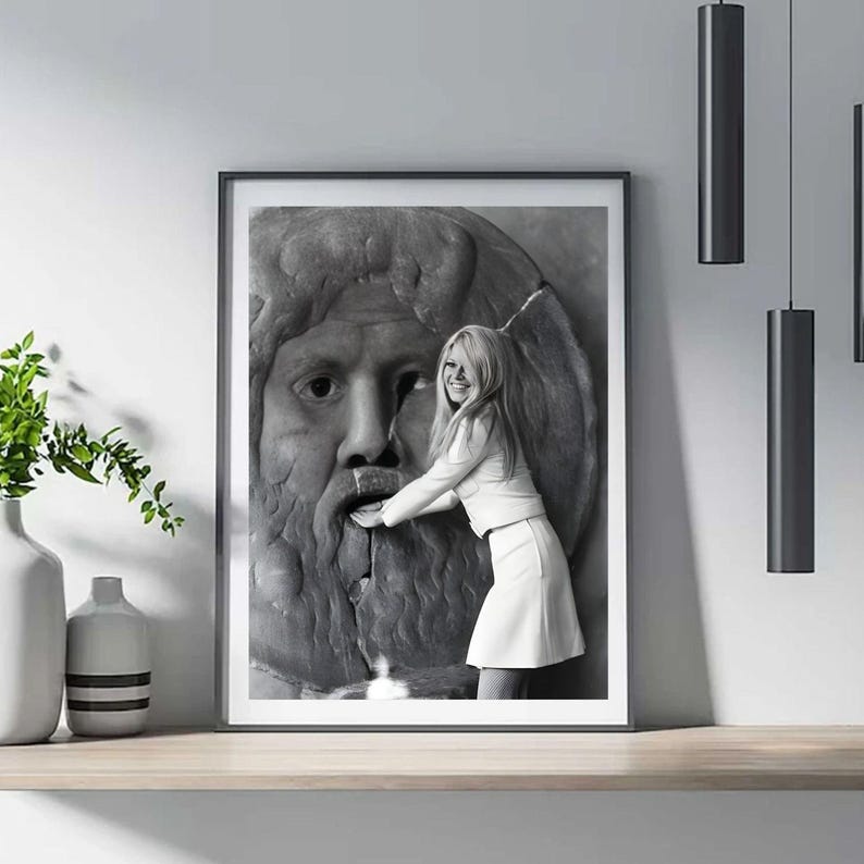 May include: Monochrome framed print showing a woman in white clothing touching a stone sculpture of a face. The print is on a wooden shelf, with a vase of greenery to the left and hanging lights.