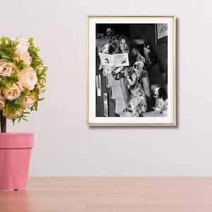 Puede incluir: Fotograf&iacute;a en blanco y negro enmarcada de una mujer leyendo un libro titulado "MOON DOG" a varios perros. La mujer est&aacute; sentada en una escalera, rodeada de perros de varias razas. La fotograf&iacute;a est&aacute; en un marco dorado.