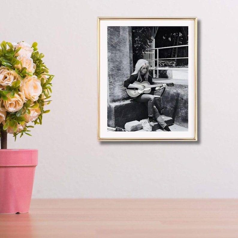 Pu&ograve; includere: Fotografia in bianco e nero incorniciata di una donna che suona una chitarra acustica. La donna &egrave; seduta all'aperto, con un top scuro e jeans. La fotografia &egrave; in una cornice dorata ed &egrave; esposta su una parete bianca.