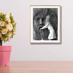 May include: A framed monochrome photograph showing a woman in a white outfit touching a stone fountain sculpture. The photograph is in a gold frame, and the background is a neutral colour.