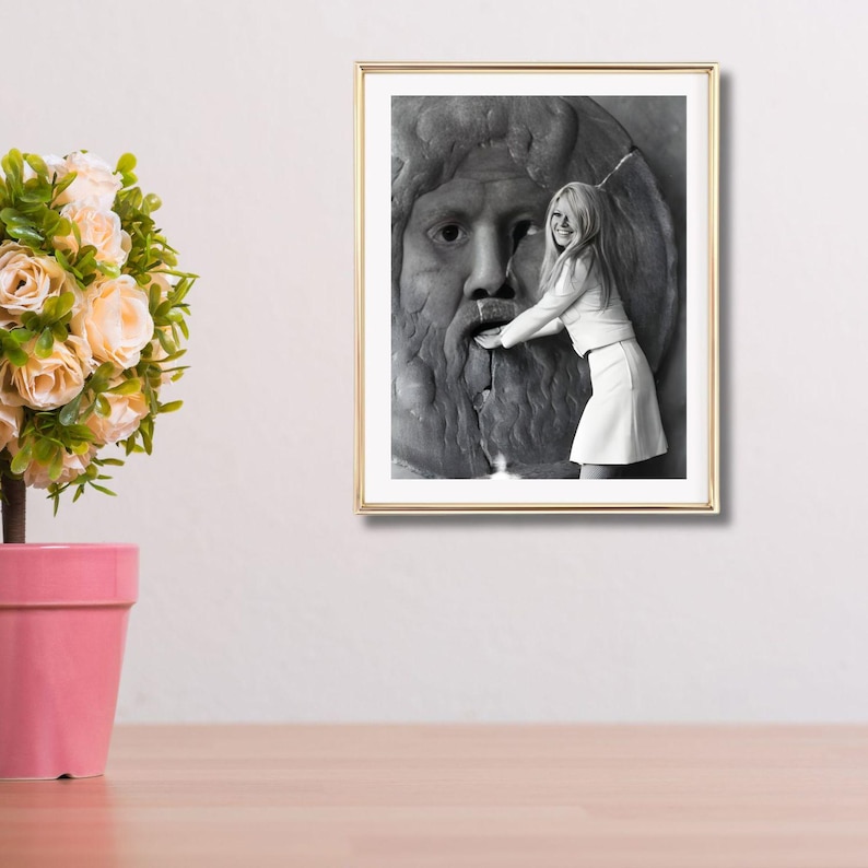 May include: A framed monochrome photograph showing a woman in a white outfit touching a stone fountain sculpture. The photograph is in a gold frame, and the background is a neutral colour.