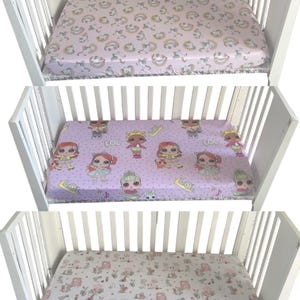 May include: Three fitted crib sheets in a white crib. The top sheet is light pink with a pattern of unicorns and rainbows. The middle sheet is lavender with LOL dolls. The bottom sheet is white with a pattern of houses and animals.