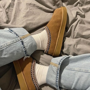 Peut inclure: Une paire de chaussons en daim marron avec une semelle beige et une bordure décorative. Les chaussures sont portées avec des chaussettes blanches et un jean bleu clair.
