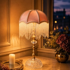 May include: A table lamp with a dome-shaped pink fabric shade featuring long, dangling beaded fringe. The lamp has a slender brass-colored metal stem, a clear faceted crystal accent, and a wide, circular, clear glass base with a diamond-cut pattern.