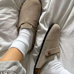 Könnte beinhalten: Hellbraune Wildlederclogs mit verstellbaren Riemen und schwarzen Sohlen. Die Clogs werden mit weißen Socken getragen. Die Schuhe liegen auf einem weißen Laken.