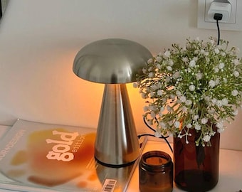 May include: A brushed metal mushroom-shaped table lamp with a conical base. The lamp is on a white surface next to a magazine with the word "gold" on the cover. A small brown jar and a vase with white flowers are also present.