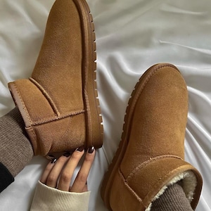 Puede incluir: Un par de botines de ante marrón con suela marrón claro. Las botas tienen un aspecto suave y texturizado y un color cálido y neutro. El interior de las botas parece estar forrado con un material blanco y esponjoso.