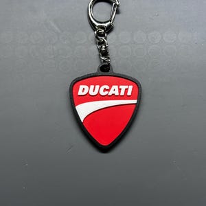 May include: A red and white Ducati logo keyring with a black border. The keyring has a silver metal ring and chain. The Ducati logo is in white text on a red shield shape.