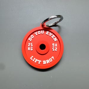 May include: A red weight plate keyring with the text "DO YOU EVEN LIFT BRO?" in white. The keyring also has the text "25 KG" on it. The weight plate is attached to a silver key ring.