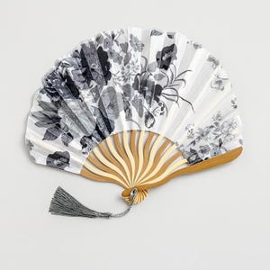 Peut inclure: Éventail blanc orné d'un motif floral noir et gris. Le manche est en bois et une houppe grise est attachée. L'éventail est ouvert, révélant le motif complexe de fleurs et de feuilles.