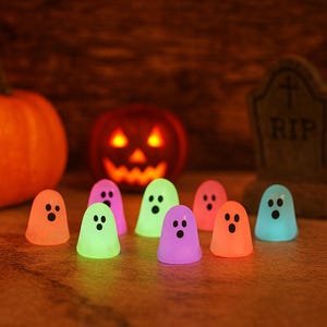 Op de afbeelding: Een Halloween-tafereel met gloeiende spookvormige lampjes in verschillende kleuren, waaronder oranje, groen en paars. Een uitgesneden pompoen en een grafsteen staan op de achtergrond, wat een spookachtige sfeer creëert. De lampjes zijn op een oppervlak geplaatst.