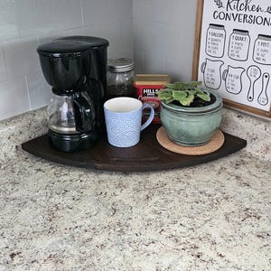 Può includere: Una scena del piano di lavoro della cucina con una caffettiera nera, una tazza blu a fantasia e una piccola pianta in vaso. Un vassoio angolare in legno contiene gli oggetti. È visibile un cartello incorniciato "Kitchen Conversions".