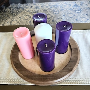 May include: A wooden candle holder with five pillar candles. The candles are pink, white, and purple. The holder has a light wood center and a dark wood rim. The candles are arranged in a circle.