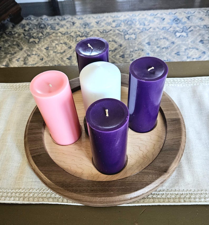 May include: A wooden candle holder with five pillar candles. The candles are pink, white, and purple. The holder has a light wood center and a dark wood rim. The candles are arranged in a circle.